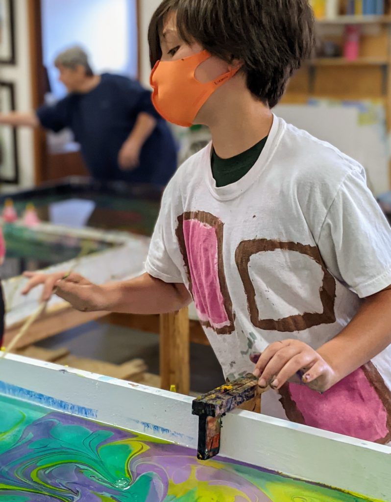 A child with an orange face mask putting paint into a marbling table.