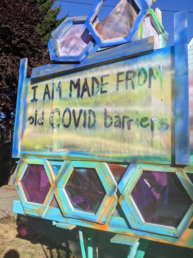 Image of a colorful sculpture made out of used covid barriers with a sign that reads "I am made of old COVID barriers."